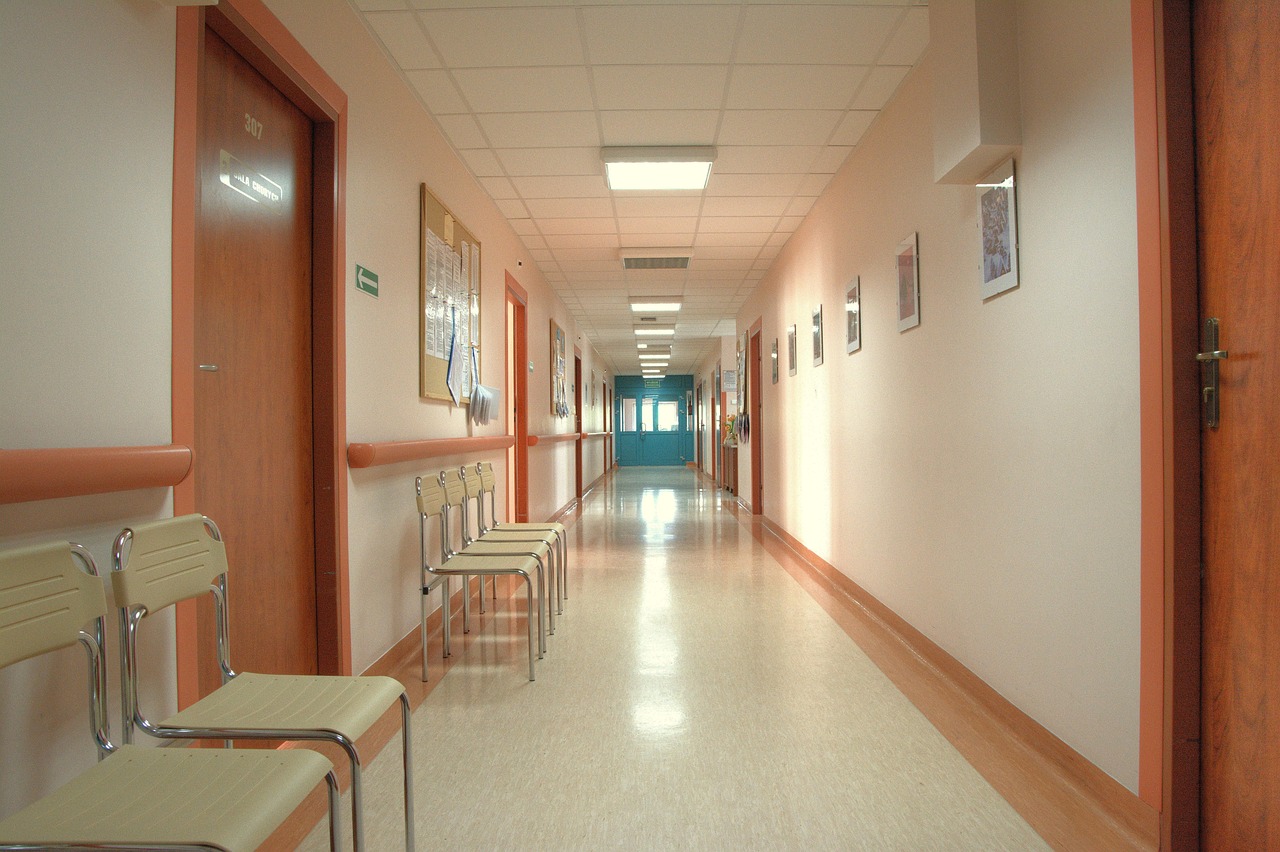 a quiet hospital corridor, with a row of chairs waiting outside closed doors