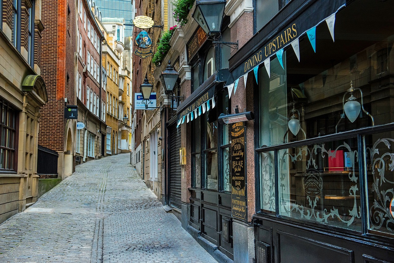 a narrow cobbled lane between old London buildings, with lanterns above the doorways