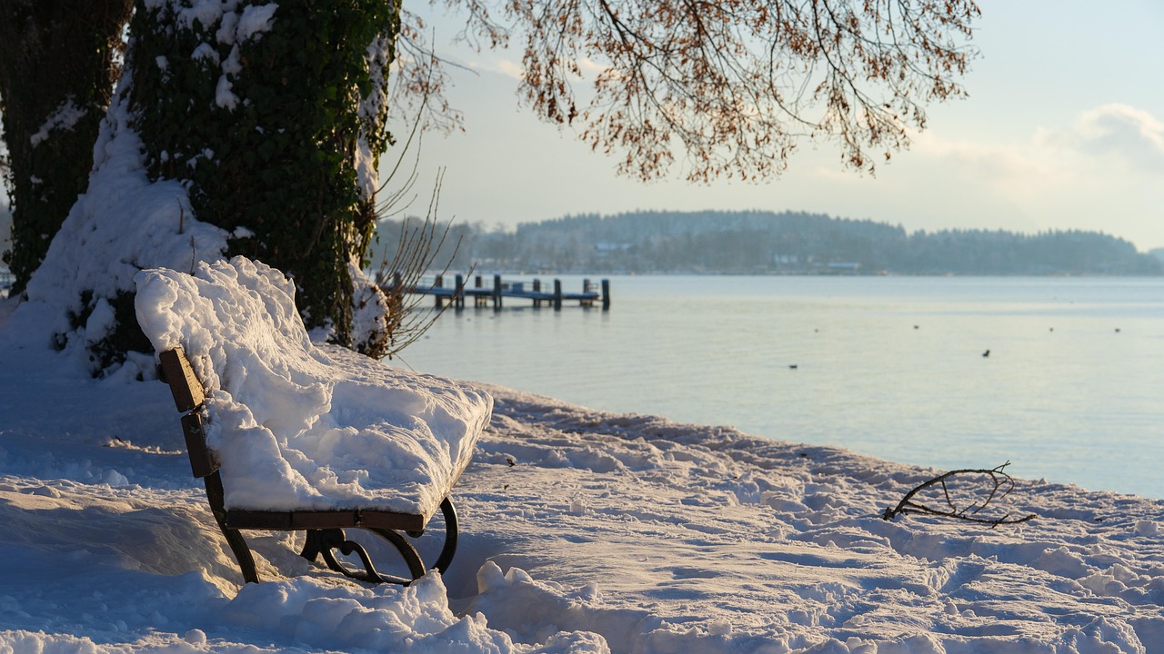 a bench heaped with snow beside a still winter lake in pale morning light