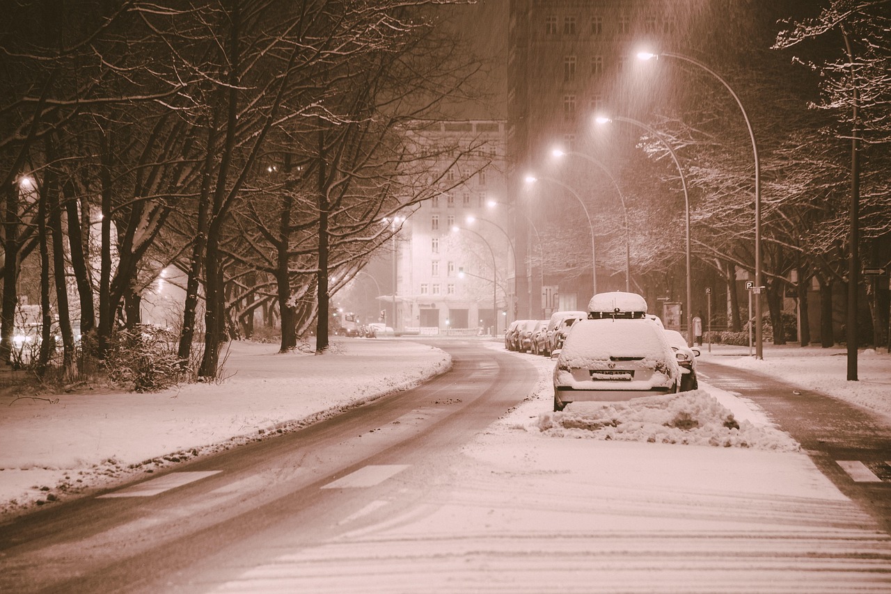 a snowy street at night, lit by streetlamps, with cars buried under snow